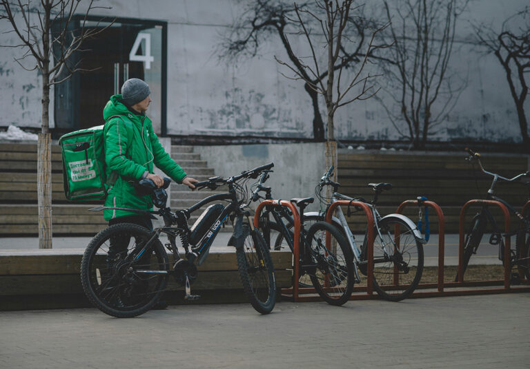 A delivery guy wearing green and carrying a bag is taking his bike from a bike rack